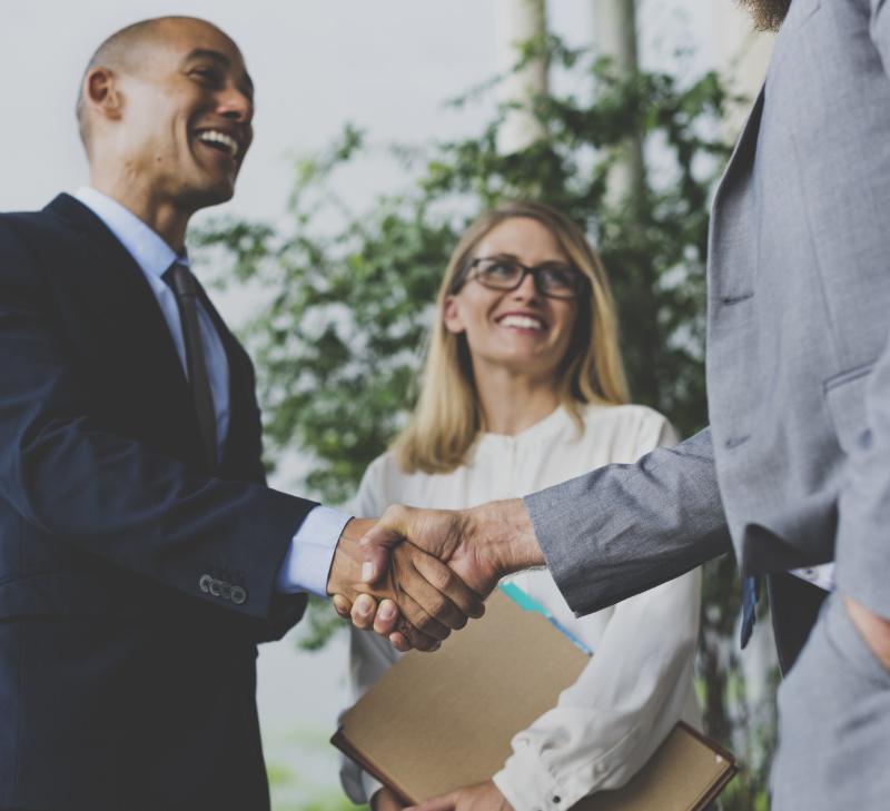 Two business men shaking hands in agreement in front of a blond smiling woman with glasses looking at one of the men
