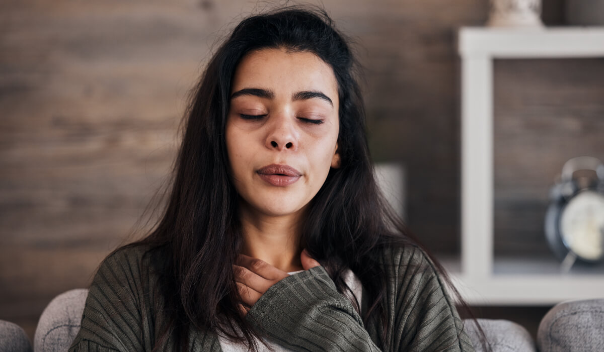 A dark haired woman with her eyes closed and hand on her throat area breathing consciously