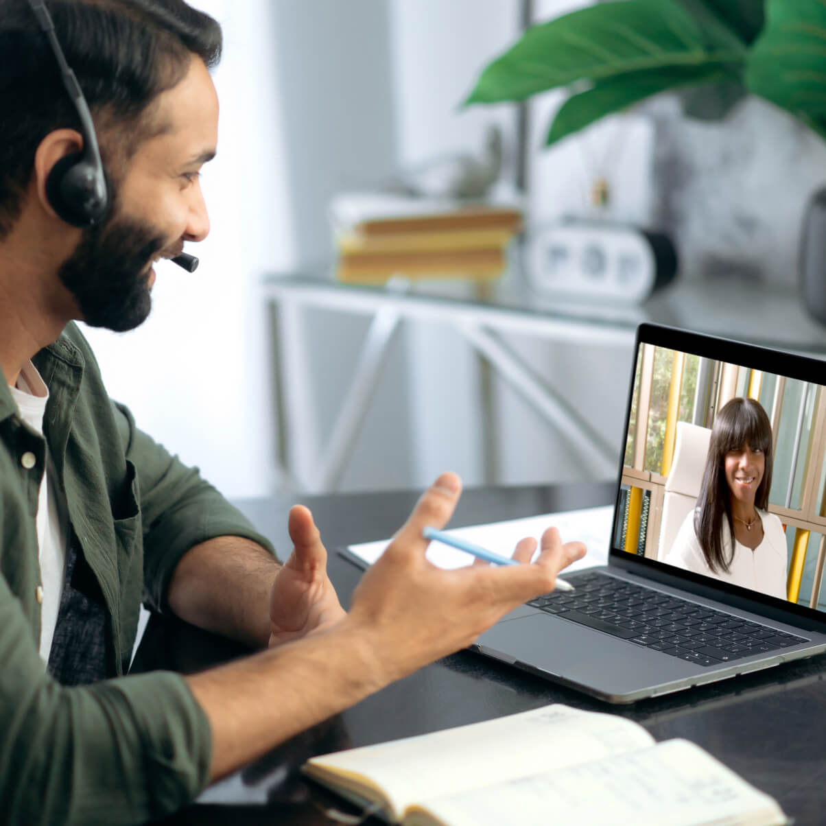 Brown haired and bearded man in office wearing a green shirt sitting at office desk with headphones taking part in a video coaching session with a black woman with long hair
